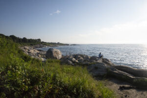 An angler fishes from a rocky shoreline as the sun rises over the water.