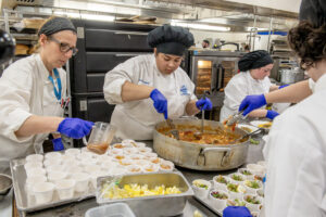Three chefs work in a kitchen stirring a pot and ladling broth into cups.
