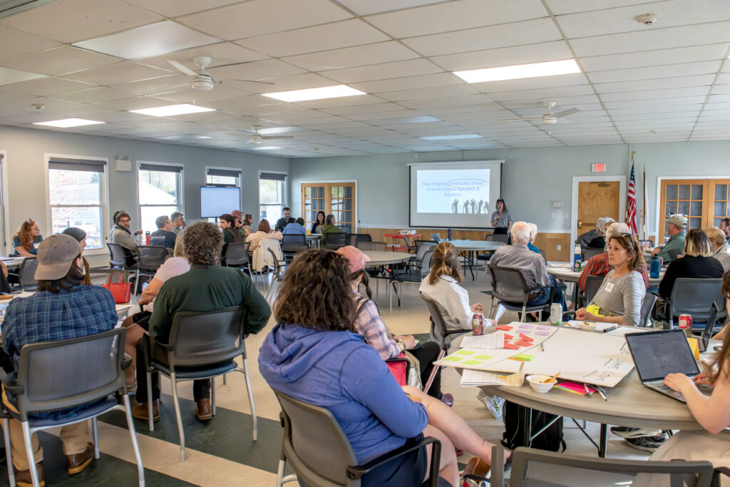 A group of people gathered around tables in a community center listens to a speaker.