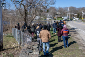 A group of students stands at the side of a road next to a fence and a stone wall