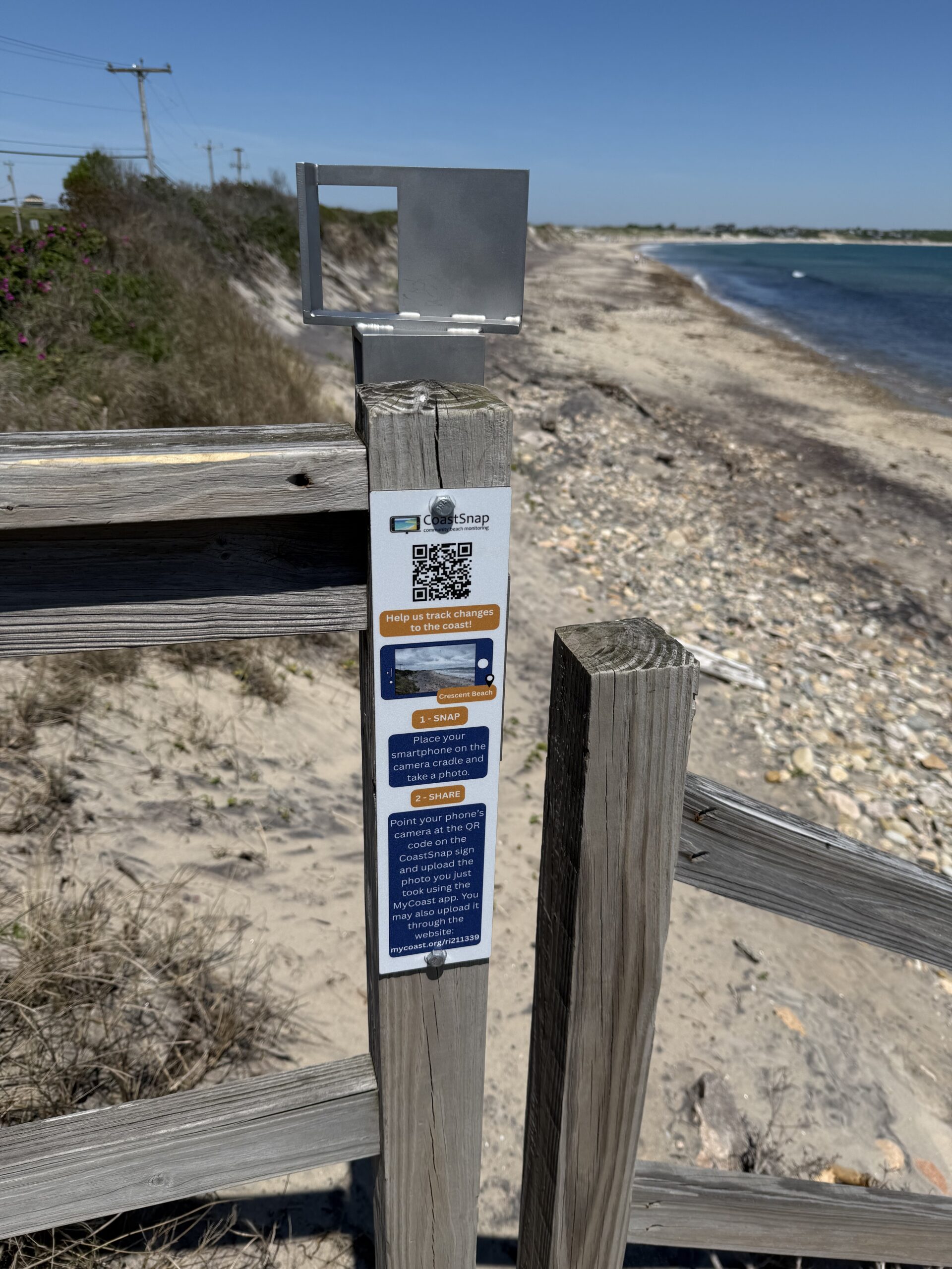 CoastSnap monitoring station installed at Crescent Beach on Block Island. A sign and smartphone mounting platform are attached to the top of a wooden beach access staircase that crosses over the dune. The station faces south toward the shoreline, where visitors can photograph the beach from a fixed location to help document coastal change over time.
