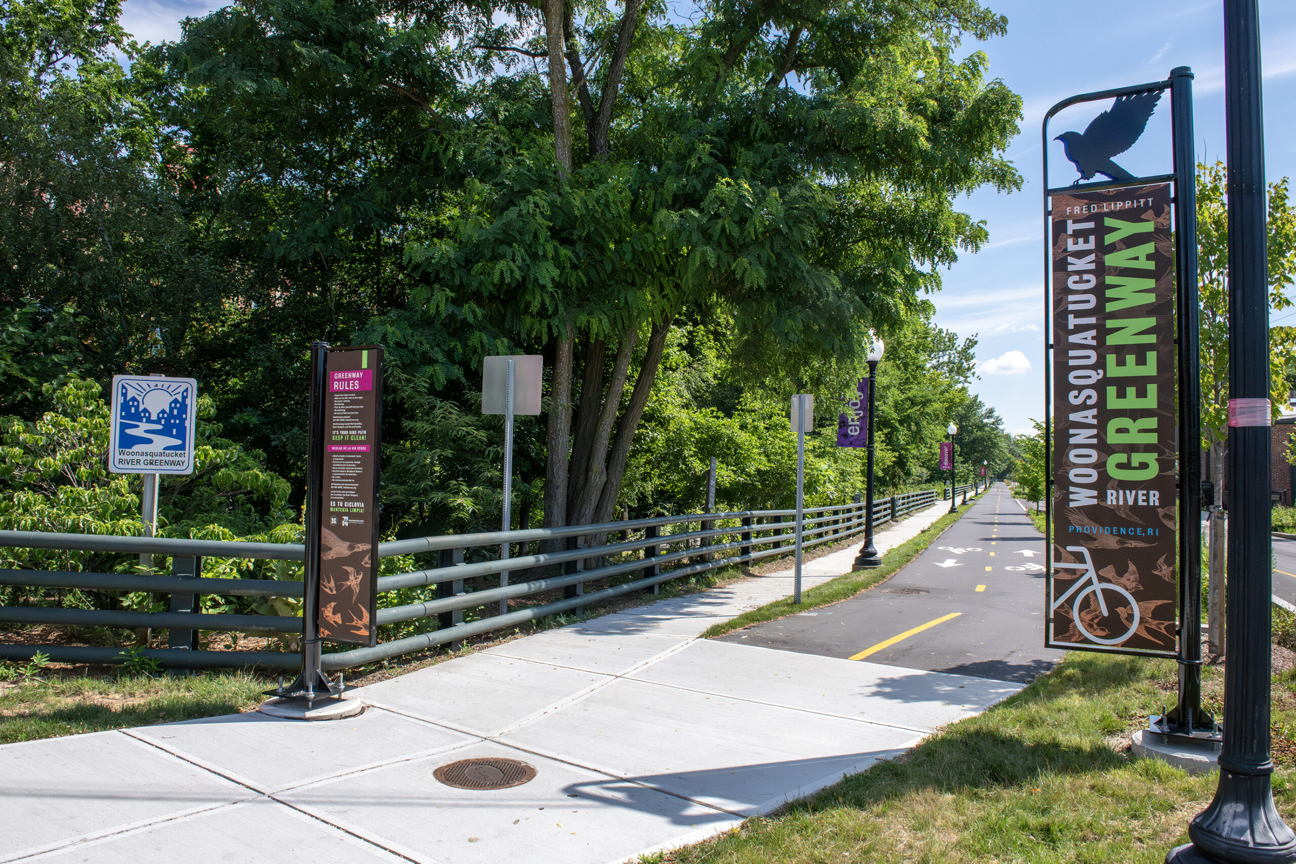 Sign reading "Woonasquatucket River Greenway" is next to a bike lane and new concrete sidewalk and shade trees