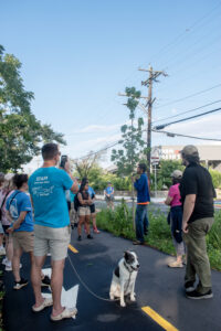 A group of people listen to a speaker at the Woonasquatucket River Greenway while a dog looks on