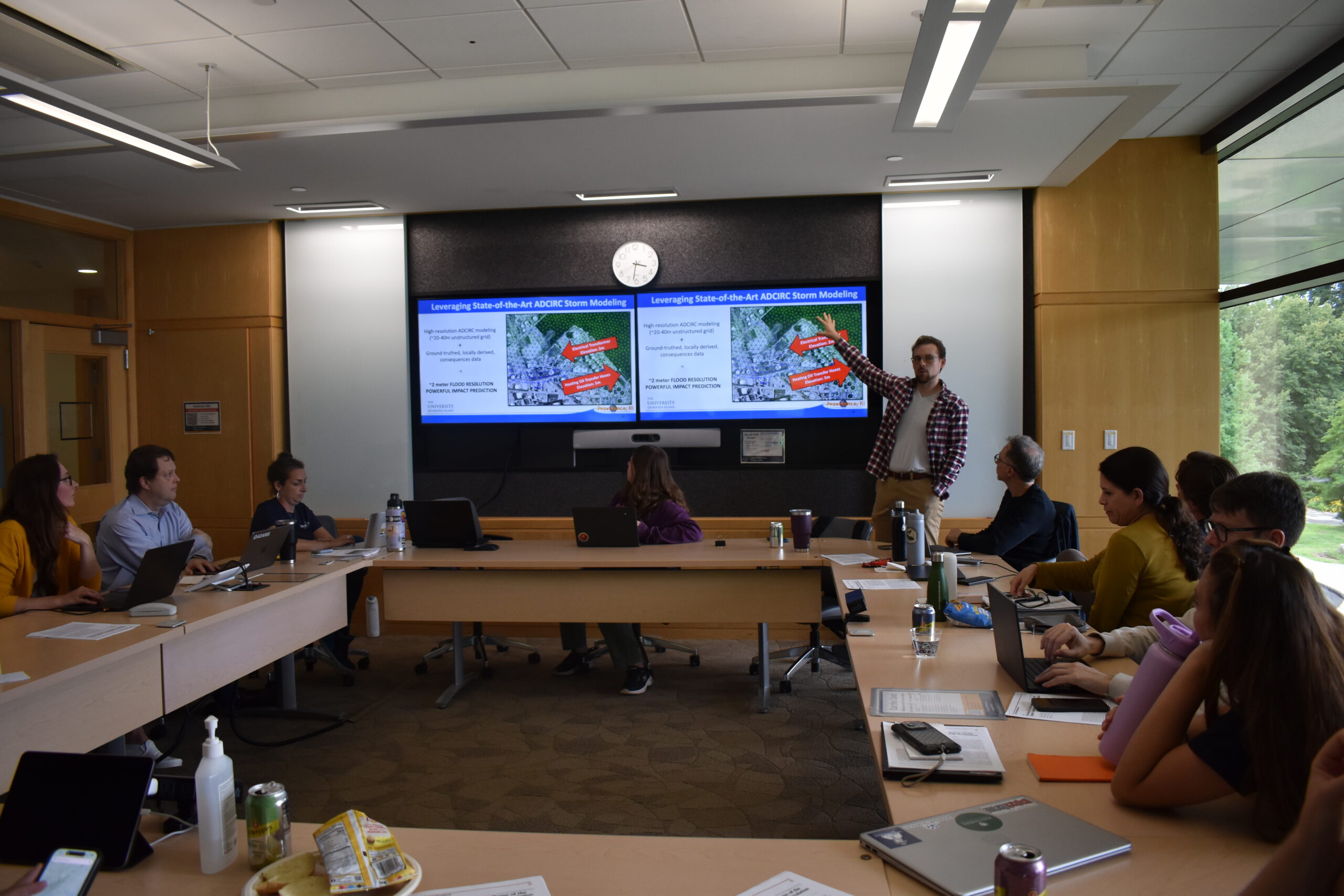 Participants in a Metcalf Institute journalism workshop sit around a conference table as a URI researcher presents the RI-CHAMP public dashboard on two screens.